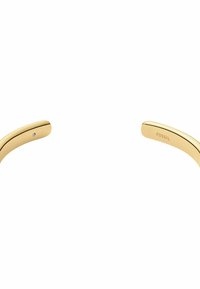 Armbandet i guldfärgad metall har släta, böjda kanter. Det har en liten ädelstensdetalj och texten "FOSSIL" ingraverad på ena sidan.