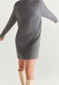 Šedé pletené šaty s vysokým límcem, dlouhými rukávy a texturovaným vzorem, sahající po střední stehno, doplněné krémovými botami po kolena.