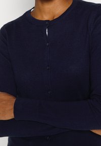 Mörkblå cardigan i lättstickad kvalitet med rund halsringning, helärm och knappstängning framtill. Mjuk textur och figurnära design.