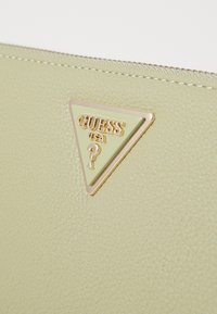 Matériau texturé vert clair avec un logo triangulaire accentué d'or portant l'inscription "GUESS USA" accompagné d'un point d'interrogation. La fermeture éclaire est visible.