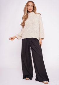 Beżowy sweter dzianinowy z wysokim kołnierzem i szerokimi rękawami, w połączeniu z czarnymi spodniami o szerokich nogawkach i jasnymi butami.