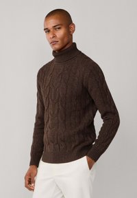 Jersey de cuello alto trenzado marrón con puños y dobladillo acanalados. La tela parece texturizada, presentando un patrón en relieve. Se lleva con pantalones de color claro.