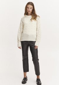 Flauschiger cremefarbener Strickpullover mit strukturiertem Muster und geripptem Saum, kombiniert mit schwarzen, cropped Lederhosen und schwarzen Slippern, vor einem neutralen Hintergrund stehend.
