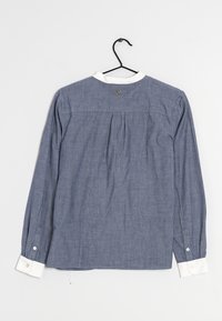 Blå chambray skjorte med hvid krave, lange ærmer og knaplukning ved håndleddene. Den har en plisseret ryg og et lille logo nær halsudskæringen.