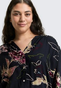 Frau mit langen, dunklen Haaren, die eine schwarze Bluse mit großen rosa, beigen und grünen Blumenmustern trägt, lächelt sanft vor einem schlichten Hintergrund.