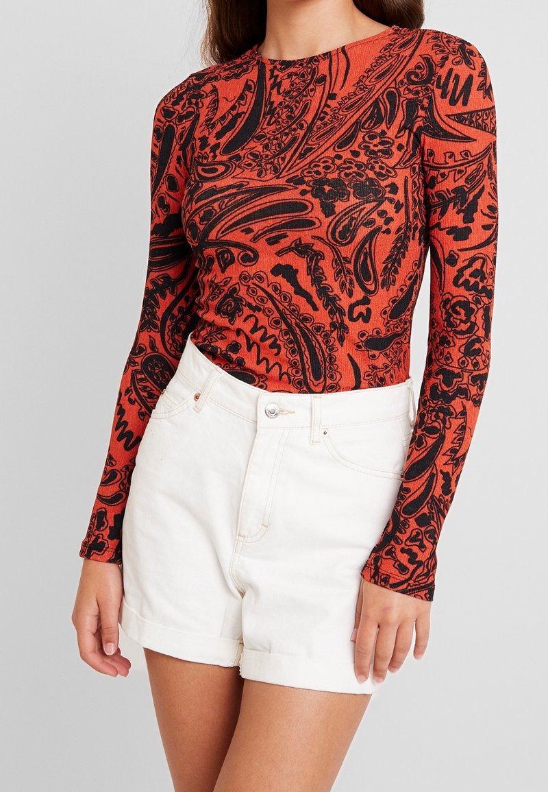 Langärmliges Oberteil in Orange mit schwarzen abstrakten Mustern, mit Rundhalsausschnitt und figurbetontem Design. Kombiniert mit weißen hochtaillierten Shorts.