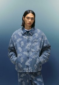 Tânăr îmbrăcat într-o jachetă de denim albastră și pantaloni asortați cu un model deschis "NYC", stând în fața unui fundal gradat albastru.