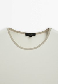 Haut en tissu côtelé crème avec une bordure beige clair autour de l'encolure ronde et une étiquette noire à l'intérieur du col.