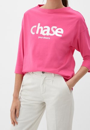 Rosa Baumwoll-T-Shirt mit lockerer Passform, weitem Ausschnitt und weißem Text, der "Verfolge deine Träume" auf der Vorderseite zeigt. Kombiniert mit leichten, lässigen Hosen.