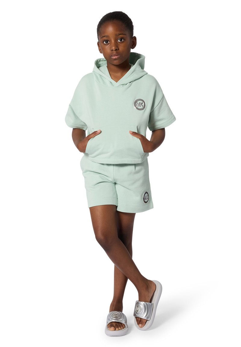 Lichtgroene hoodie met korte mouwen, voorkantzakken en logo. Bijpassende korte broek, gladde textuur. Grijze sandalen met een metalen band.