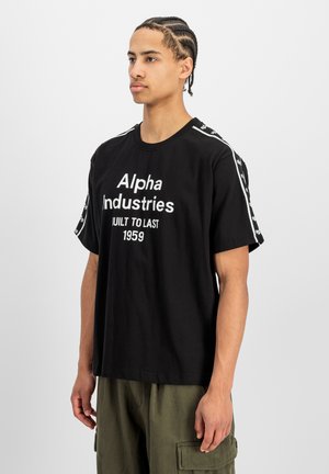 Junger Mann mit geflochtenem Haar, der ein schwarzes Alpha Industries T-Shirt und olivgrüne Cargohosen trägt, steht vor einem schlichten grauen Hintergrund.