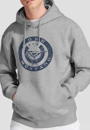 Mann trägt grauen Kapuzenpullover mit blauem, rundem Ford Mustang-Logo mit laufendem Pferd und Sternen auf der Vorderseite, Hände in den Taschen des Kapuzenpullovers.