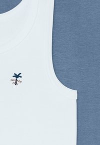 Biała koszulka na ramiączkach z prążkowanym dekoltem i małą grafiką palmowego drzewa, obok gładkiej niebieskiej tkaniny bawełnianej o aksamitnej fakturze.