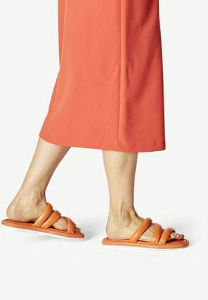 Oranje schuiflippers met drie dikke banden en een platte zool, gemaakt van glad materiaal, gedragen met een lange koraalkleurige rok.