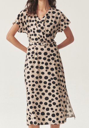 Beige midi-kjole med sorte uregelmæssige polkaprikker, V-hals, korte ærmer, bælte i taljen og sideslidser, båret af en stående kvinde.