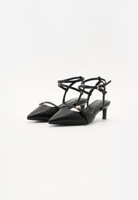 Talons hauts en cuir noir à dos nu avec un embout pointu, présentant des bretelles croisées et de petits accents dorés. Design de talon bas et effilé.