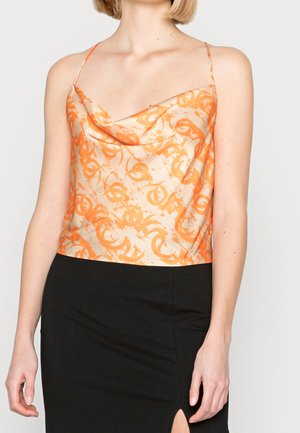 Vrouw draagt een mouwloos oranje zijden top met patroon en dunne bandjes, gecombineerd met een zwarte rok met een vooraan gespleten zoom.