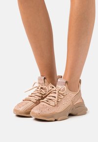 Sneakers atletiche beige con tomaia texturizzata, impreziositi da strass, dotati di lacci e un'etichetta con logo sul lato.