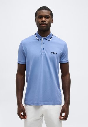 Uomo che indossa una polo azzurro chiaro con il logo "BOSS" sul petto, bordo scuro sul colletto e pantaloni bianchi su sfondo bianco uniforme.