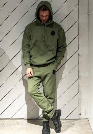 Bluza z kapturem i spodnie dresowe w kolorze oliwkowej zieleni, wykonane z miękkiego materiału, z przednią kieszenią i naszywką z logo na nogawce. Czarne buty.