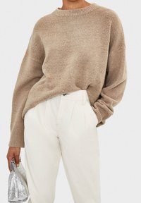 Beige knitted sweater with a round neckline and relaxed fit. Paired with white high-waisted trousers. Silver textured handbag held in hand.