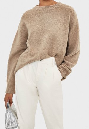 Beige strikket sweater med rund halsudskæring og afslappet pasform. Kombinationen med hvide bukser med høj talje. Sølvfarvet tekstureret håndtaske holdt i hånden.