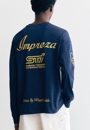 Osoba nosící tmavě modrou košili s dlouhým rukávem s žlutým nápisem „Impreza STI Subaru Tecnica International“ na zádech a bílými kalhotami.