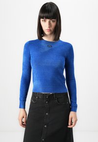 Vrouw met steil donker haar die een aansluitend felblauw langemouwen shirt en een hoge zwarte knooprok draagt, staat tegen een witte achtergrond.