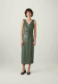 Žalia blizgučių suknelė su gilia V formos iškirpte, be rankovių, su raukšlių juosmens detale ir midi ilgiu, derinama su juodais bateliais su blokuotu kulnu.