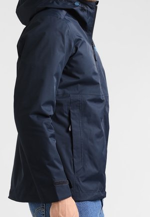 Mand iført en mørkeblå hættetrøje med lynlås og velcrobånd ved ærmerne, vist fra siden mod en hvid baggrund.