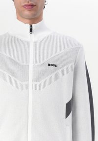 Bela zip-up jopica z teksturiranim vzorcem, ki ima kontrastni črn poudarek in logotip. Visok ovratnik in lahka pletenina.