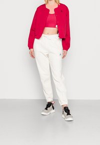 Red bomber jacket, pink crop top, white joggers with elastic cuffs, and black and gray sneakers with red accents. Logo visible on clothing.