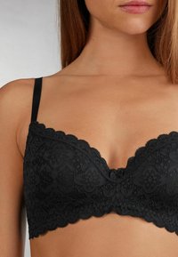 Černá krajková podprsenka s květinovými vzory, krajkovými okraji, nastavitelnými ramínky a hladkým texture; navržena pro těsné přilnutí.