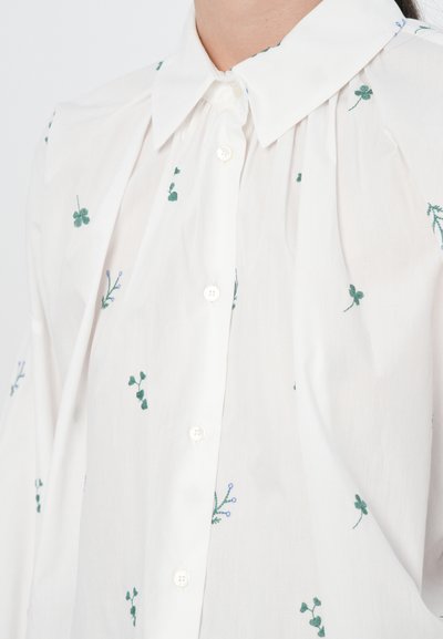 Chemise blanche à boutons avec broderie florale verte et accents bleus, dotée d'un col classique et de détails froncés sur les épaules.