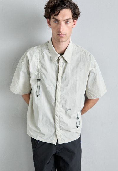Chemise à manches courtes beige clair avec un col, dotée de cordons de serrage ajustables et de détails de couture minimalistes.