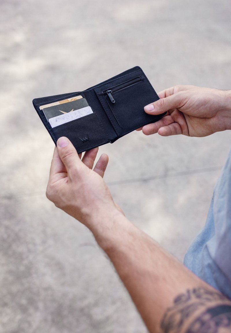 Black wallet made of fabric, opened to show card slots and a zippered pocket, held with two hands against a blurred background.
