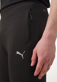Melnas sporta bikses, kas izgatavotas no mīksta auduma, ar rāvējslēdzēja kabatu un baltu Puma logo sānos. Redzamas teksturētas sānu paneļi.