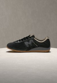 Baskets basses noires avec des lacets noirs, une semelle grise et beige, et un branding "Marc Jacobs" sur le panneau latéral, sur une surface neutre.