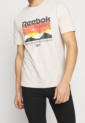 Man draagt een beige Reebok Classic t-shirt met een kleurrijke bergafbeelding en de tekst "Adventure Happens Everywhere," gecombineerd met een zwarte broek.