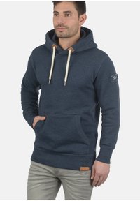 Marinblå hoodie med kängurufickor, krämfärgade snoddar och bruna läderdetaljer. Har ribbade ärmslut och nederkant, tillverkad av mjuk tyg.