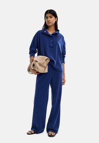 Niebieski sweter z małym kołnierzykiem i zatrzaskami, w zestawieniu z pasującymi spodniami o szerokich nogawkach. Beżowa torba i płaskie sandały dopełniają stylizację.