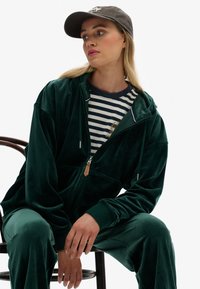 Grön sammetszip-tröja med sidofickor, i kombination med en marinblå och vit randig t-shirt. Modellen bär en grå keps. Slät textur, avslappnad passform.