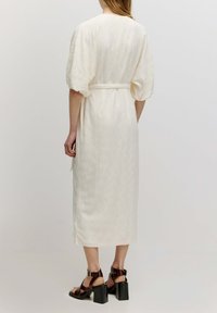 Langes weißes Kleid mit strukturiertem Muster, kurzen Puffärmeln und einem Bindegürtel in der Taille; kombiniert mit schwarzen Blockabsatzsandalen.
