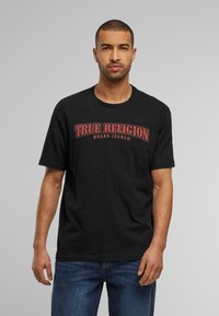 Camiseta de algodón negra con un logo rojo de "TRUE RELIGION BRAND JEANS" en la parte delantera. Cuello redondo, mangas cortas, corte estándar.