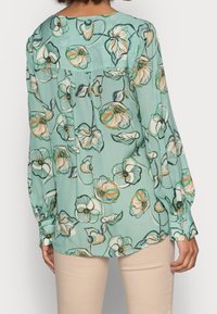 Mujer vistiendo una blusa azul claro con un patrón floral naranja y blanco, de mangas largas, y pantalones beiges, vista desde atrás.