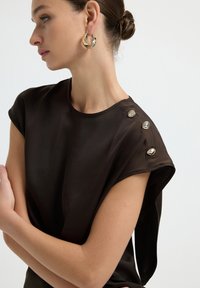 Top corto marrone a maniche corte con una texture liscia, dotato di bottoni decorativi sulla spalla e un design drappeggiato sulla manica. Orecchini a cerchio dorati.