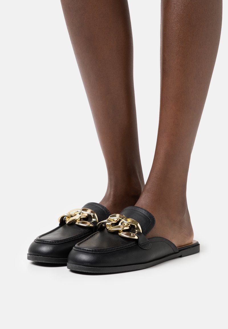 Mocassins noirs en cuir à enfiler avec un grand détail de chaîne dorée, portés pieds nus sur un fond clair uni.