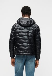 Άνδρας με γυαλιστερό μαύρο μπουφάν με κουκούλα και μπλε τζιν, εμφανίζεται από πίσω σε απλό λευκό φόντο.