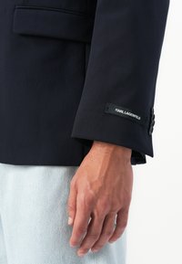 Vyriška ranka šalia tamsiai mėlynos švarkelio rankovės su Karl Lagerfeld etiketė ir juodais mygtukais, derinama su šviesiai mėlynomis džinsinėmis kelnėmis.