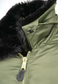 Alyvuogių žalios spalvos striukė su juodos dirbtinės kailio apykakle, auksinės spalvos užtrauktuku ir juodos spalvos užtrauktuko laikikliu, pagaminta iš lygaus, tvirto audinio.